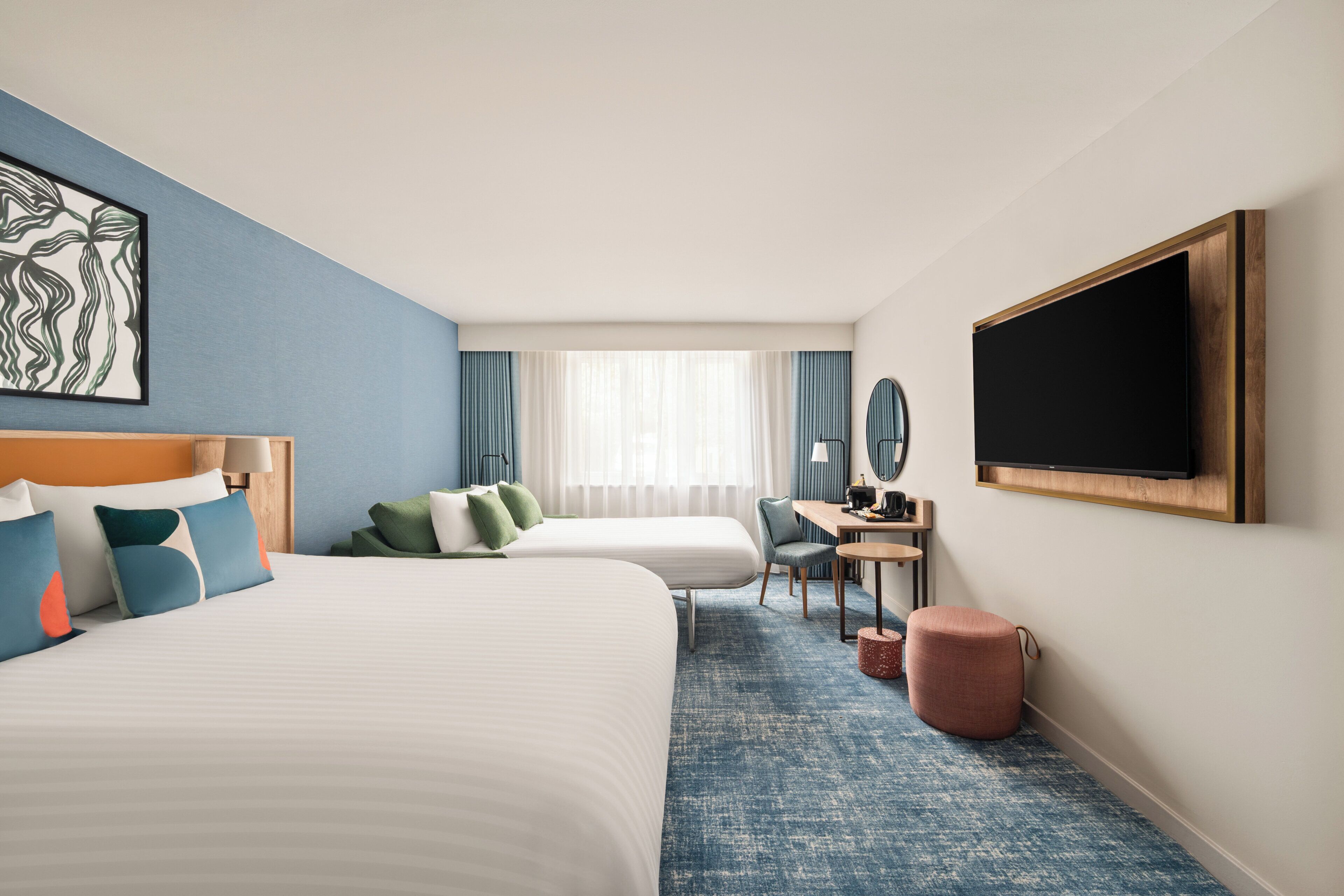 Habitación de hotel con dos camas, un escritorio, una silla, un televisor y un ventanal con cortinas.