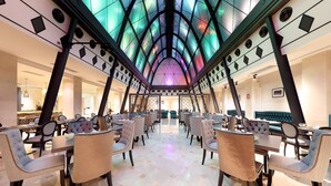 A spacious dining area with high ceilings and a modern chandelier. Tables are arranged in a U-shape, surrounded by upholstered chairs.