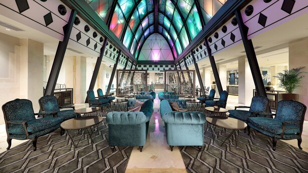 A spacious lobby with a high ceiling, featuring a geometric glass structure and seating areas with upholstered chairs and tables.