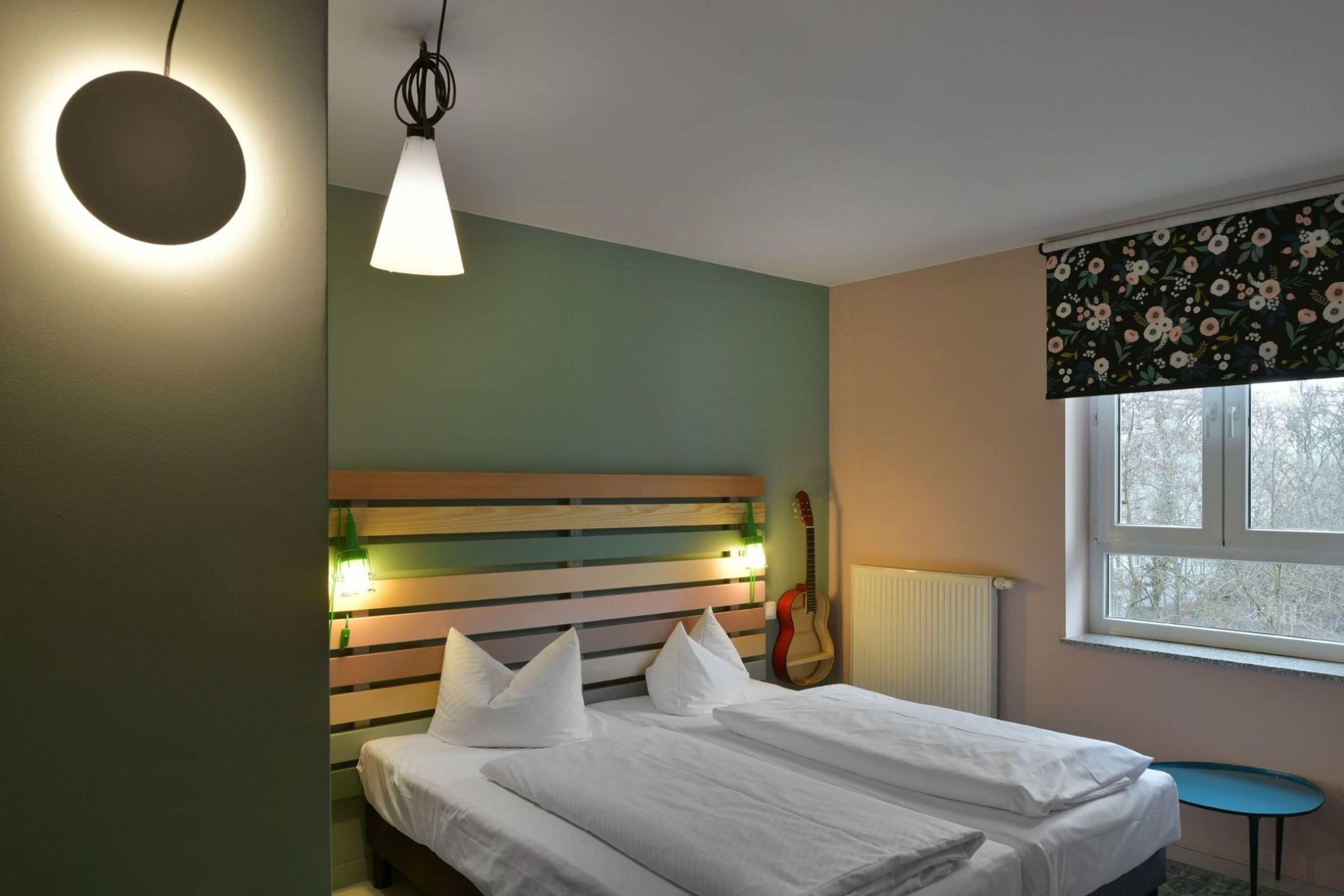 Une chambre à coucher avec un lit, une guitare, une petite table et une fenêtre ornée d’un rideau à motifs floraux.
