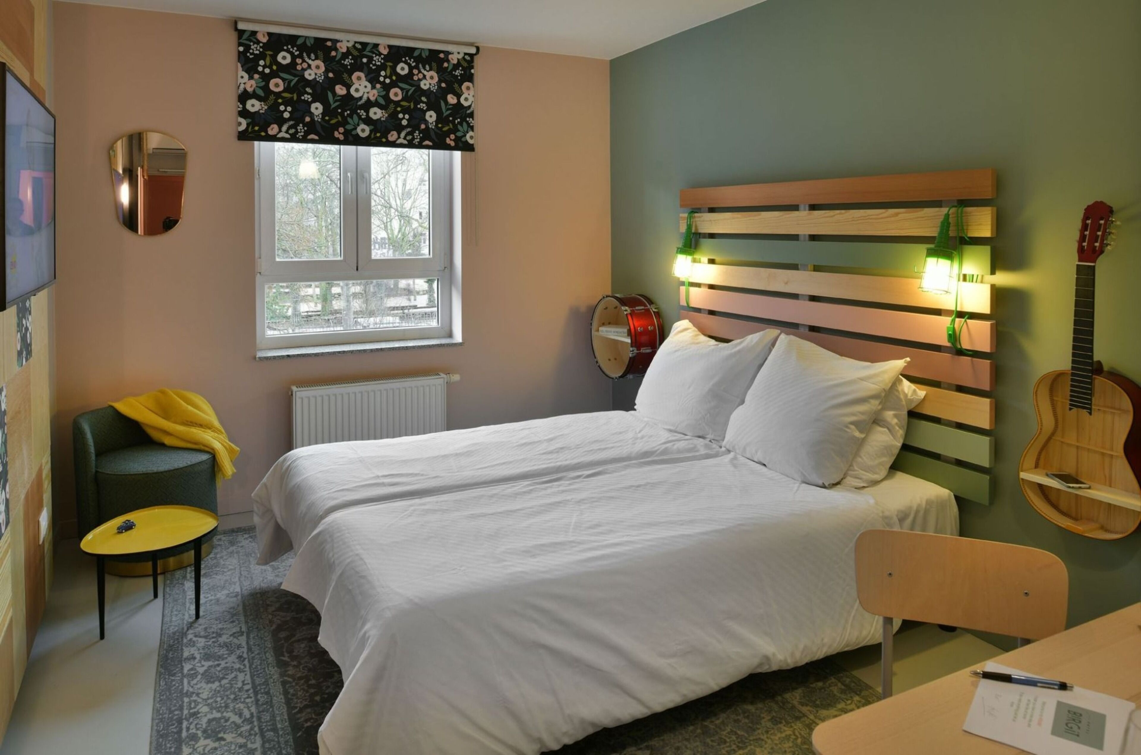 Une chambre à coucher avec un lit, une chaise, une table, une guitare et une fenêtre avec un radiateur.