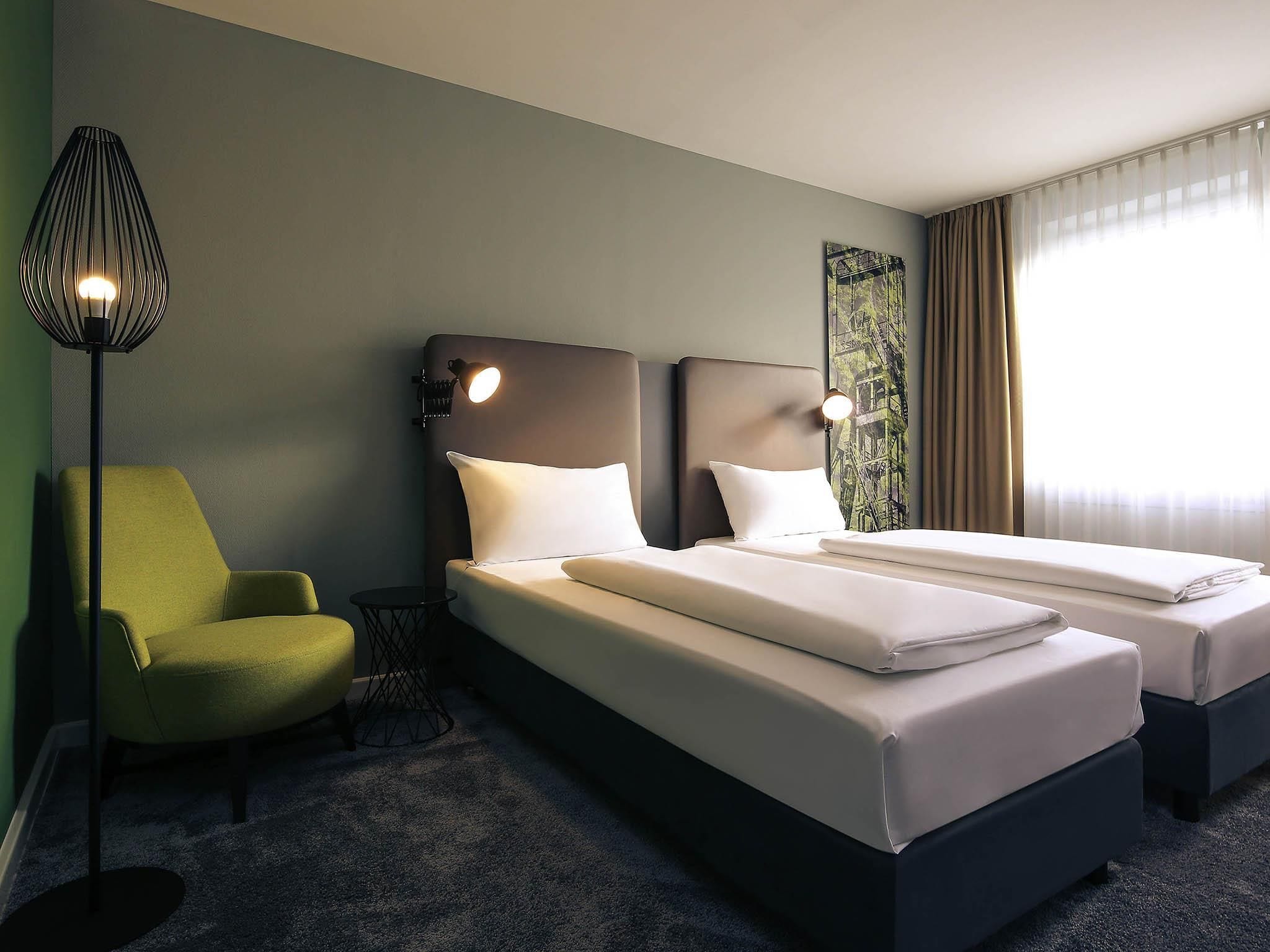 Habitación de hotel con dos camas, un sillón verde, una lámpara de pie y una ventana con cortinas.