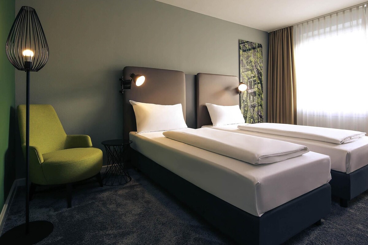 A hotel room with two beds, a green armchair, a floor lamp, and a window with curtains.