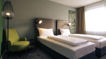 Habitación de hotel con dos camas, un sillón verde, una lámpara de pie y una ventana con cortinas.
