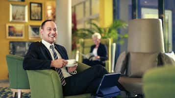 A man sitting on a green armchair with a laptop and a cup, in a lobby with framed pictures on the wall and a person in the background.