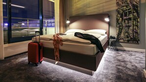 Habitación de hotel moderna con una cama grande, una maleta y vistas de un paisaje urbano a través de amplias ventanas.