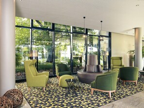 Un área de descanso moderna con ventanas amplias, sillones tapizados en verde, una zona de estar central y un piso con estampado.