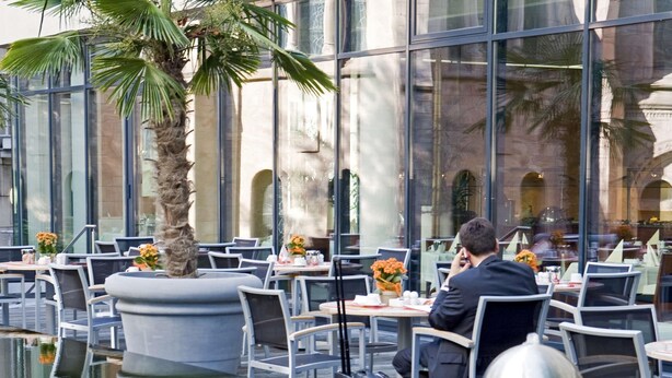 Un espacio al aire libre para comer con mesas y sillas, una palmera y un edificio con grandes ventanales al fondo.