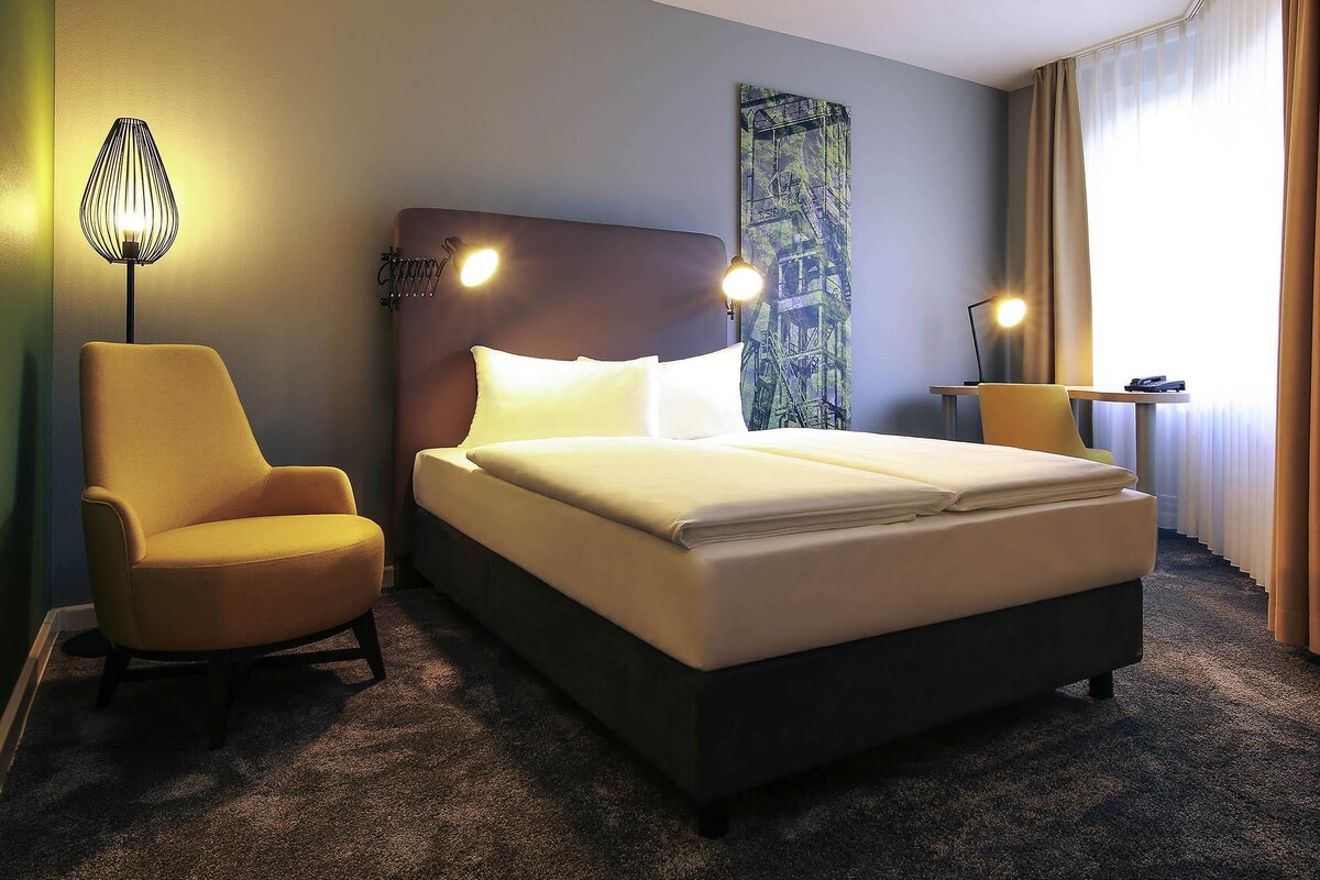 A hotel room with a bed, a chair, a lamp, and a painting on the wall.