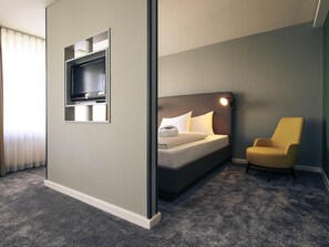 Una habitación de hotel con una cama, un televisor montado en la pared, una silla y suelo alfombrado.