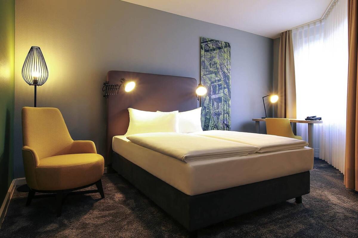 Quarto de hotel com cama, uma cadeira, um abajur e um quadro na parede.