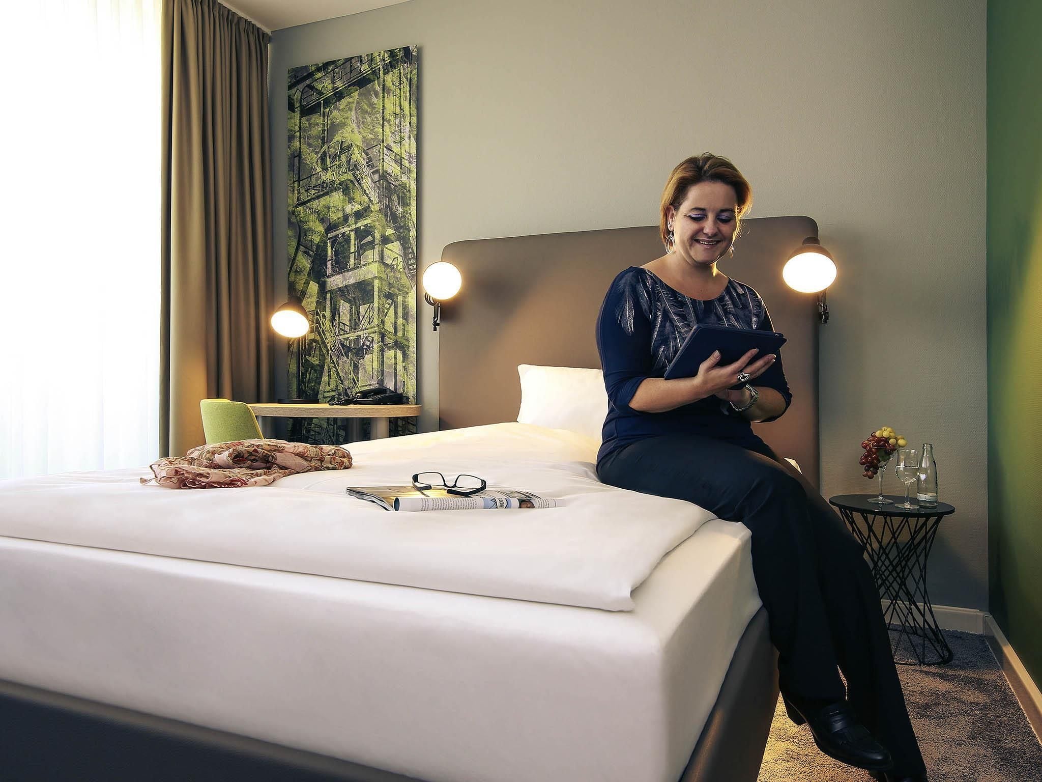 A hotel room with a bed, a bedside table with a lamp, a wall art piece, and a small table with a vase and glasses.
