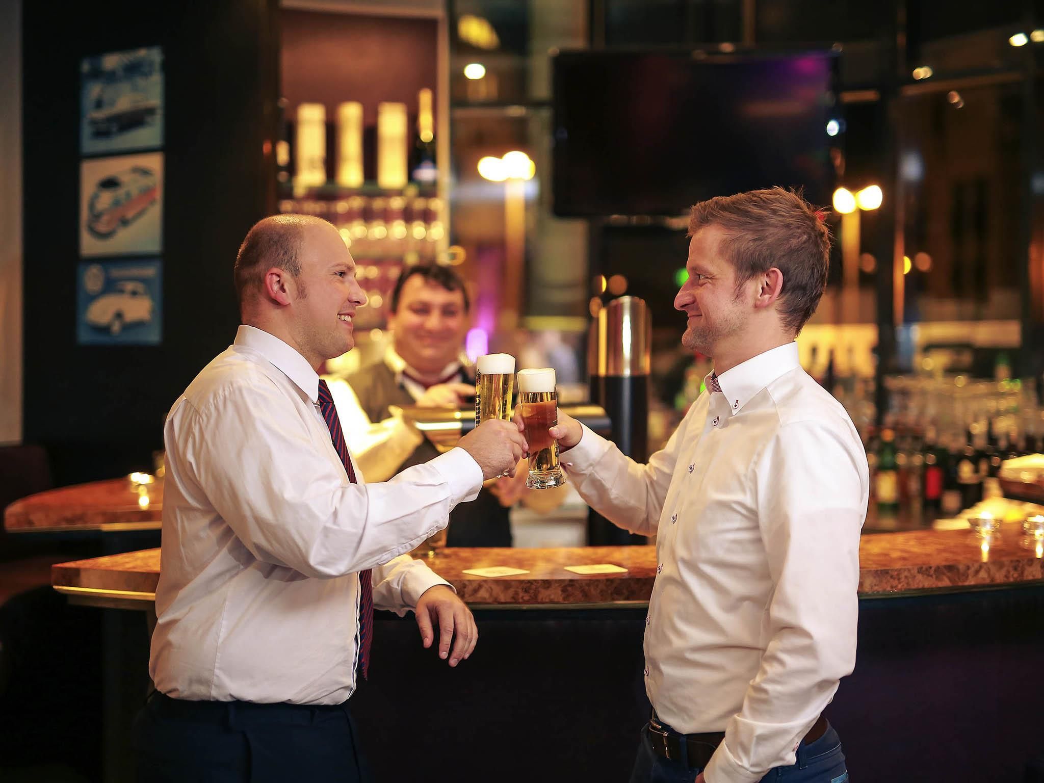 Dos hombres brindando con vasos de cerveza en un bar, con barra y estantes llenos de botellas.