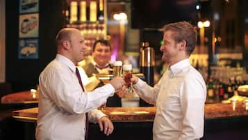 Dos hombres brindando con vasos de cerveza en un bar, con barra y estantes llenos de botellas.