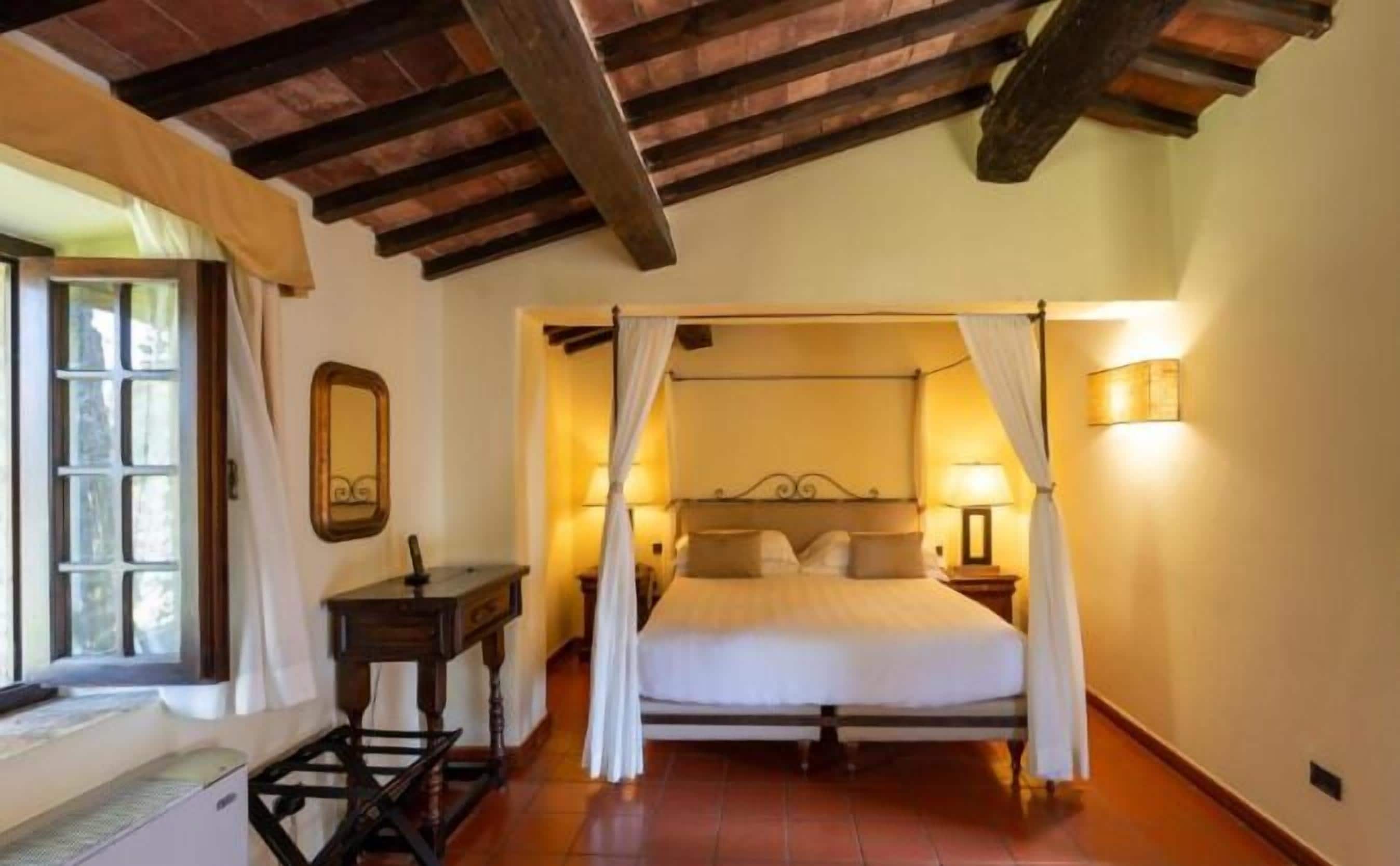 A bedroom with a four-poster bed, a bedside table, a mirror, and a window with curtains.
