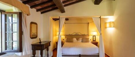 A bedroom with a four-poster bed, a bedside table, a mirror, and a window with curtains.