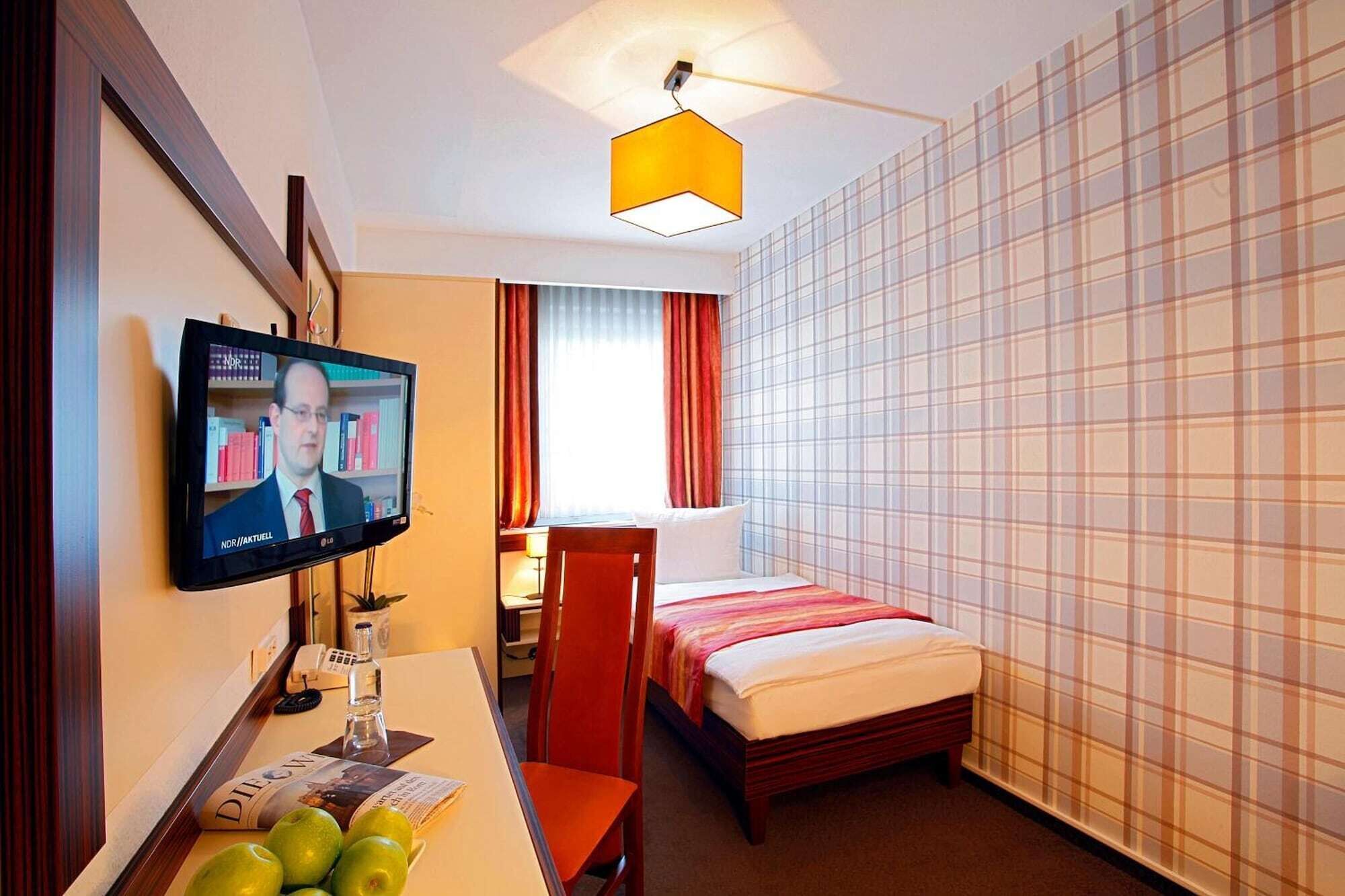 Ein Hotelzimmer mit Bett, Schreibtisch mit Computer, Fernseher und Fenster mit Vorhängen.