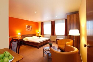 Ett hotellrum med en säng, ett skrivbord, en stol, en lampa och ett bord med äpplen.