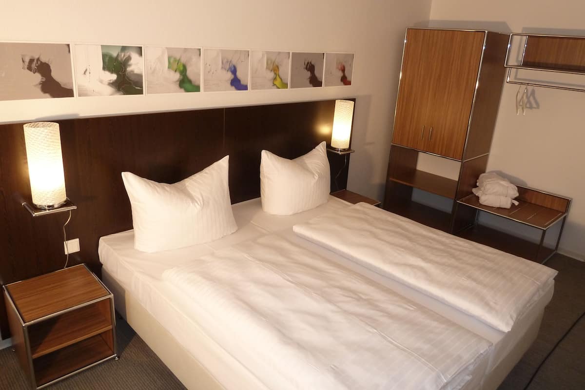 Un lit bien fait, avec du linge de lit blanc et des oreillers, une table de chevet avec une lampe et une armoire avec un miroir.
