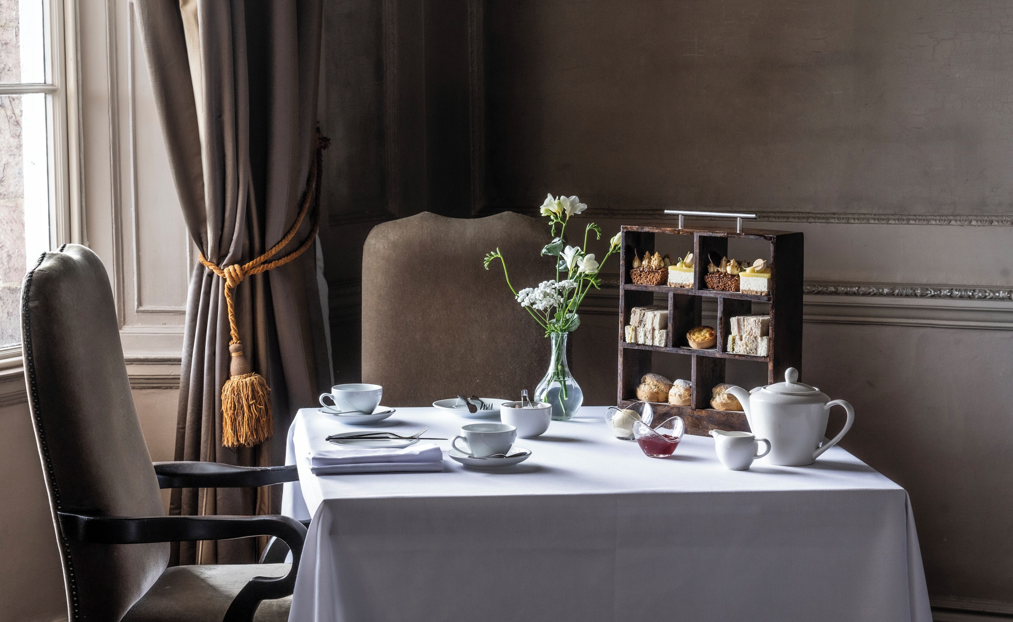 A dining table with a white tablecloth, a vase with flowers, a tea set, and a dessert display.
