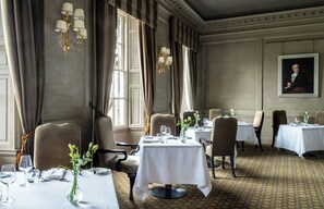 A formal dining room with tables set for service, elegant chandeliers, and a classical painting on the wall.