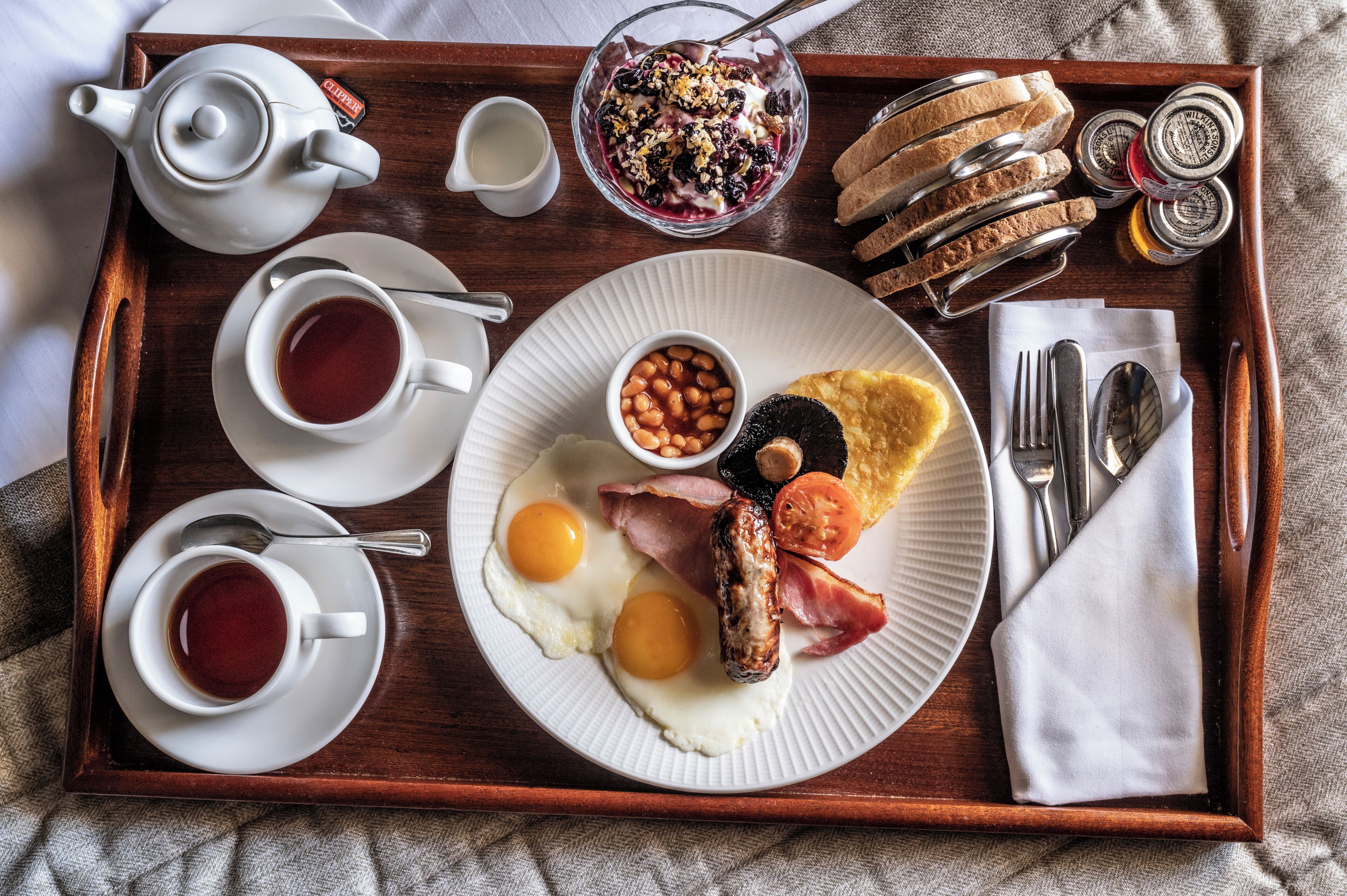 A breakfast tray with eggs, sausage, bacon, toast, jam, and a teapot.