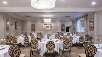 A banquet hall with round tables, chairs, chandeliers, and framed pictures on the walls.