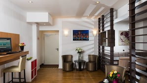 A modern hotel room with a wooden desk, two leather chairs, a small round table with a vase of flowers, and a wall-mounted TV. - Leonardo Hotel Frankfurt City Center (Frankfurt)