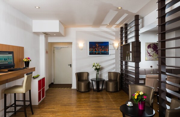 A modern hotel room with a wooden desk, two leather chairs, a small round table with a vase of flowers, and a wall-mounted TV. - Leonardo Hotel Frankfurt City Center (Frankfurt)