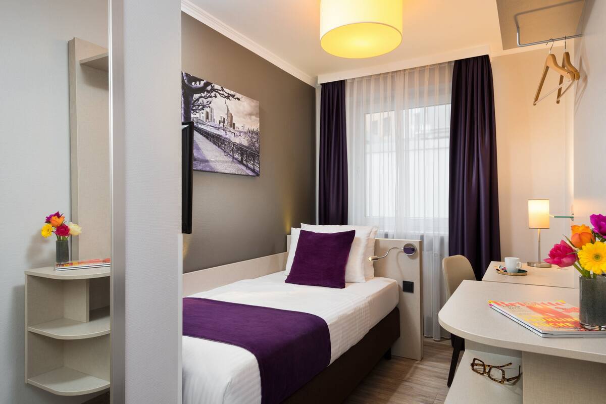 A hotel room with a bed, a desk, a chair, a window with curtains, a wall-mounted lamp, and a painting.
