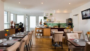 A modern hotel dining area with a central island, multiple seating options, and a kitchen in the background. - Leonardo Hotel Frankfurt City Center (Frankfurt)