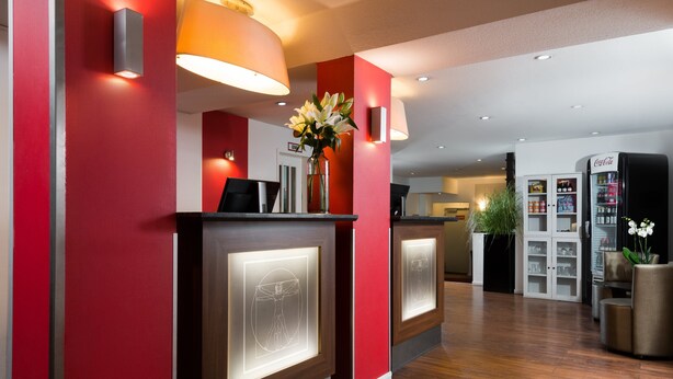 A reception area with a red wall, a reception desk, a Coca-Cola refrigerator, and a potted plant. - Leonardo Hotel Frankfurt City Center (Frankfurt)