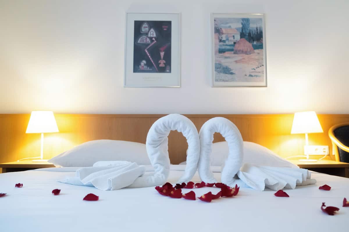 Un lit soigneusement fait, avec des serviettes disposées en forme de cœur, des pétales de rose éparpillés sur le lit et deux œuvres d’art encadrées accrochées au mur.