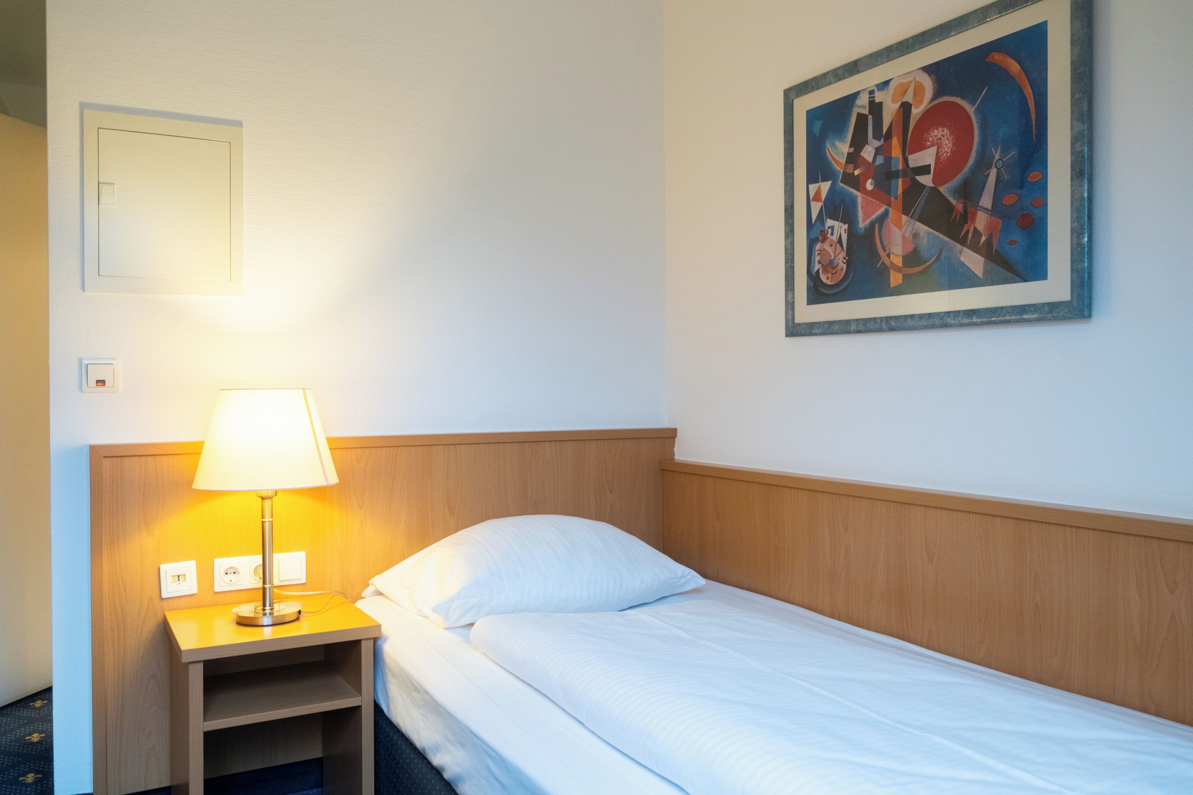 A hotel room with a bed, a nightstand with a lamp, a painting on the wall, and a power outlet.