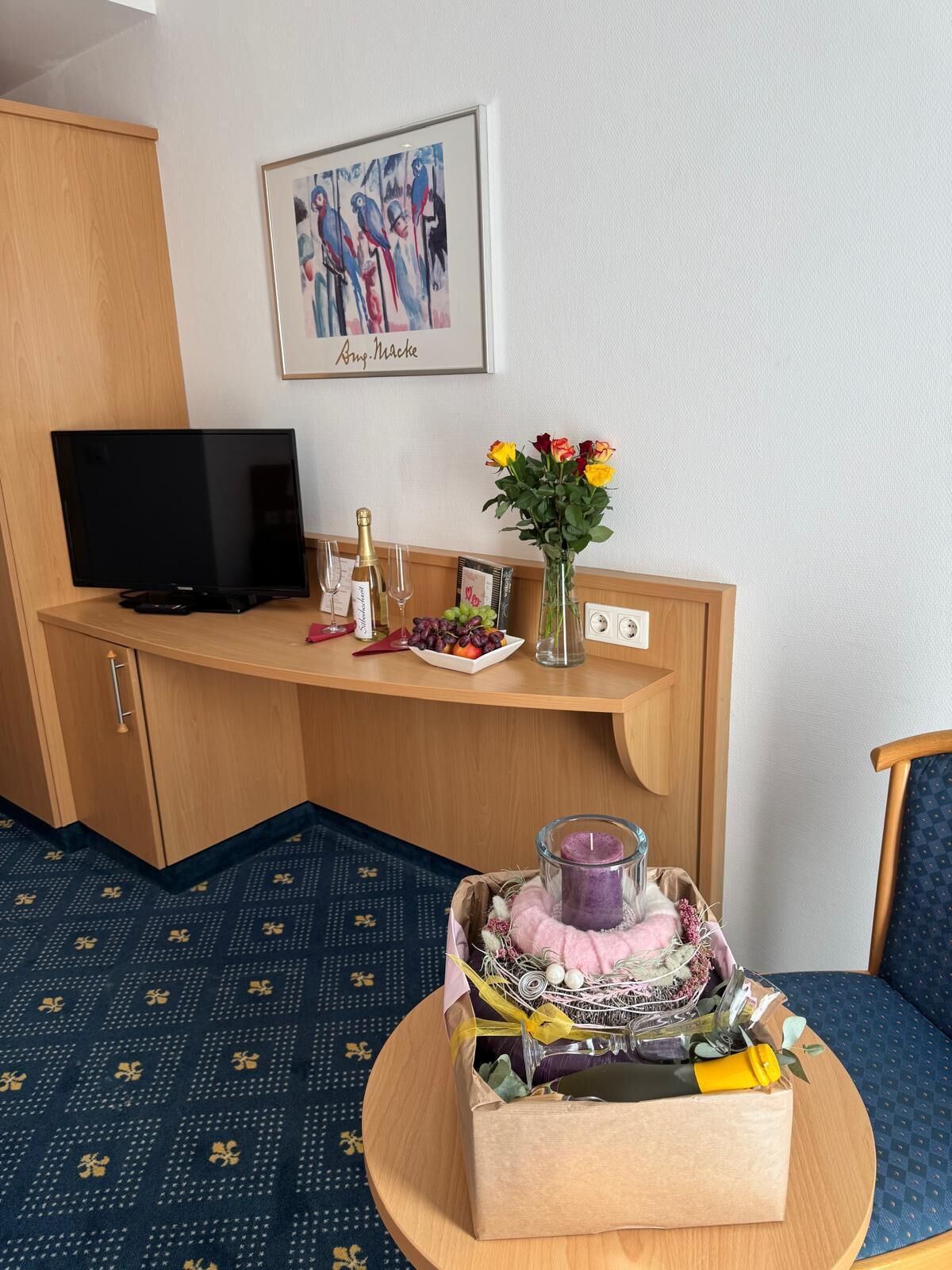 A hotel room with a TV, a small table with a candle, a vase with flowers, and a framed picture on the wall.