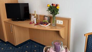A hotel room with a TV, a small table with a candle, a vase with flowers, and a framed picture on the wall.