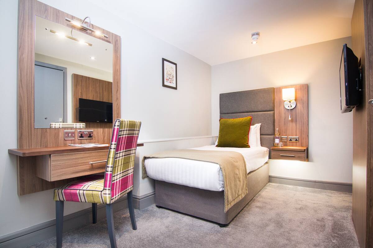 A hotel room with a bed, a desk with a chair, a television, and a bedside table with a lamp.