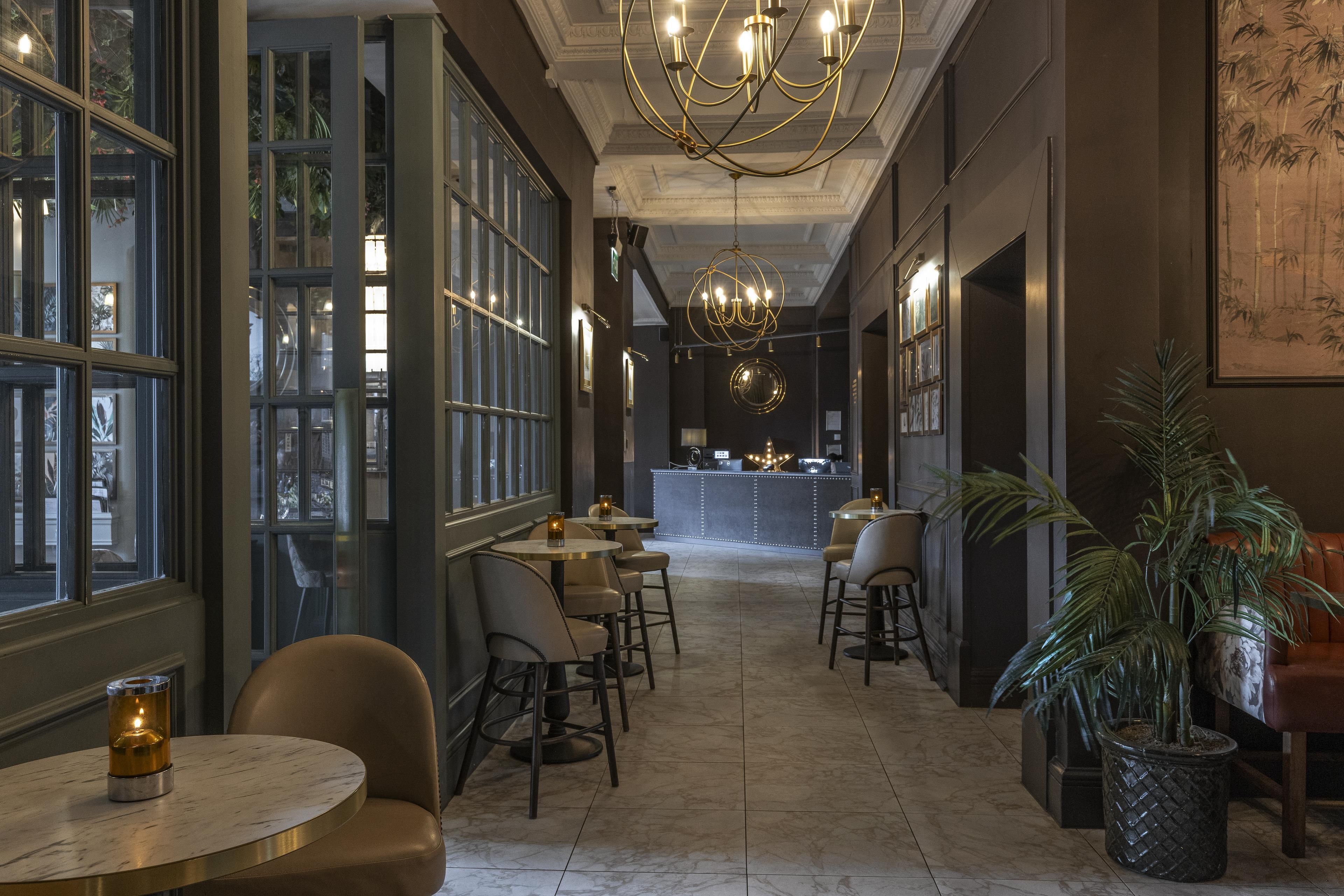 Un couloir d’hôtel avec des tables rondes, des chaises, un coin bar et un grand lustre.