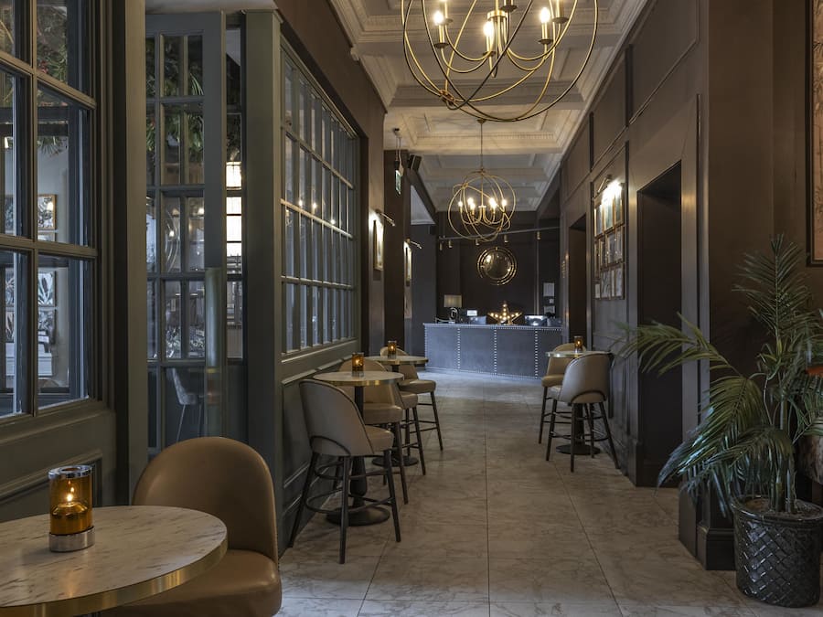 Un pasillo de hotel con mesas redondas, sillas, una zona de bar y una gran lámpara de araña.