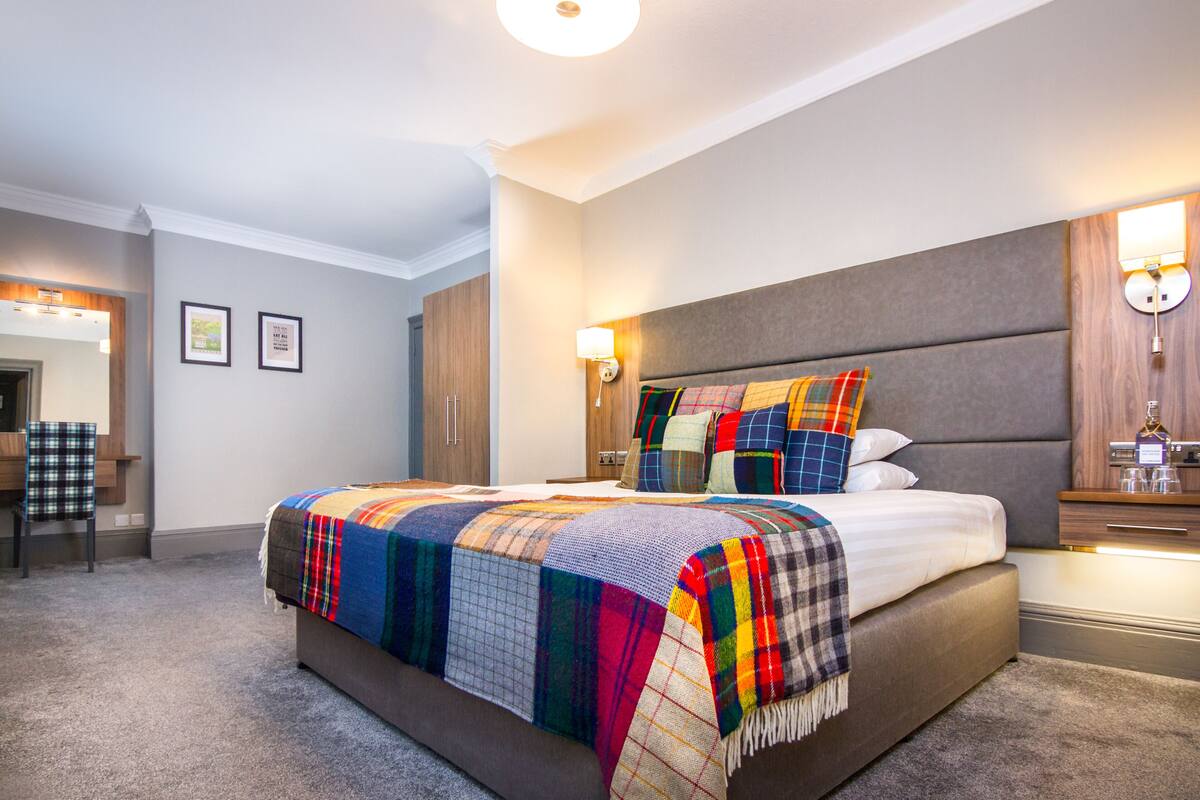 A hotel room with a large bed, a nightstand with a lamp, a chair, and a wooden wardrobe.