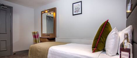 A neatly arranged hotel room with a bed, a bedside table with a bottle, a mirror, and a framed picture on the wall.