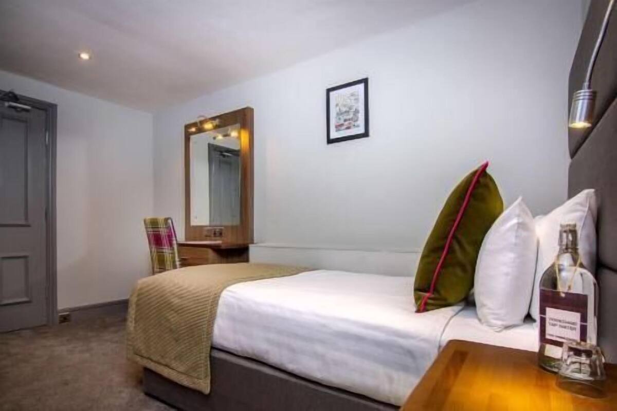 A hotel room with a bed, a bedside table with a bottle, a mirror, and a framed picture on the wall.