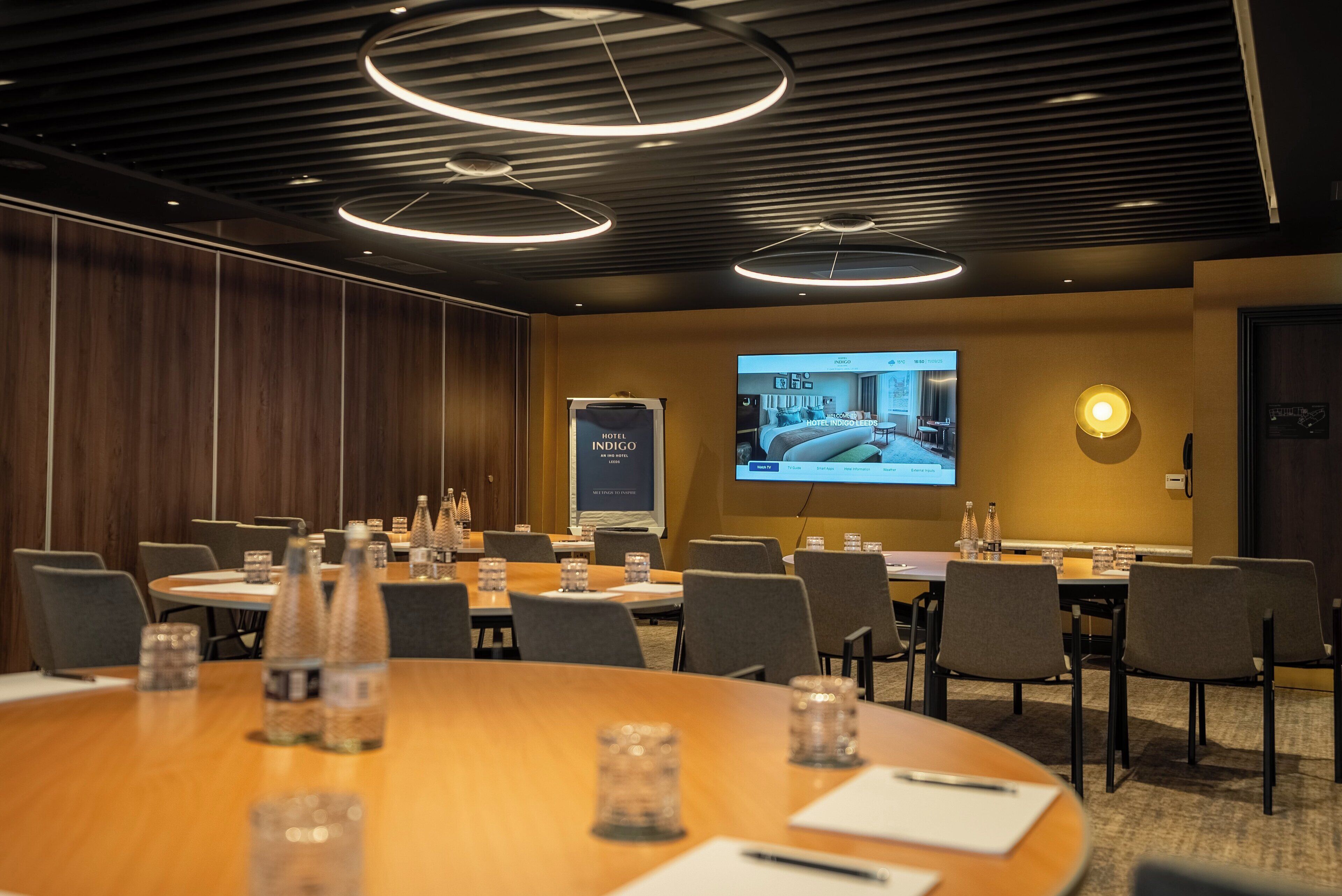 A conference room with a large screen displaying a hotel interior, round tables with water bottles, and a wooden panel wall.