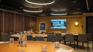 A conference room with a large screen displaying a hotel interior, round tables with water bottles, and a wooden panel wall.