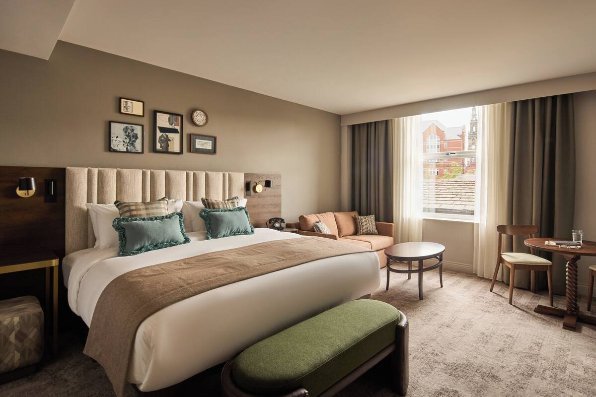 Una habitación de hotel con una cama grande, un sofá, una mesita y una ventana con cortinas.