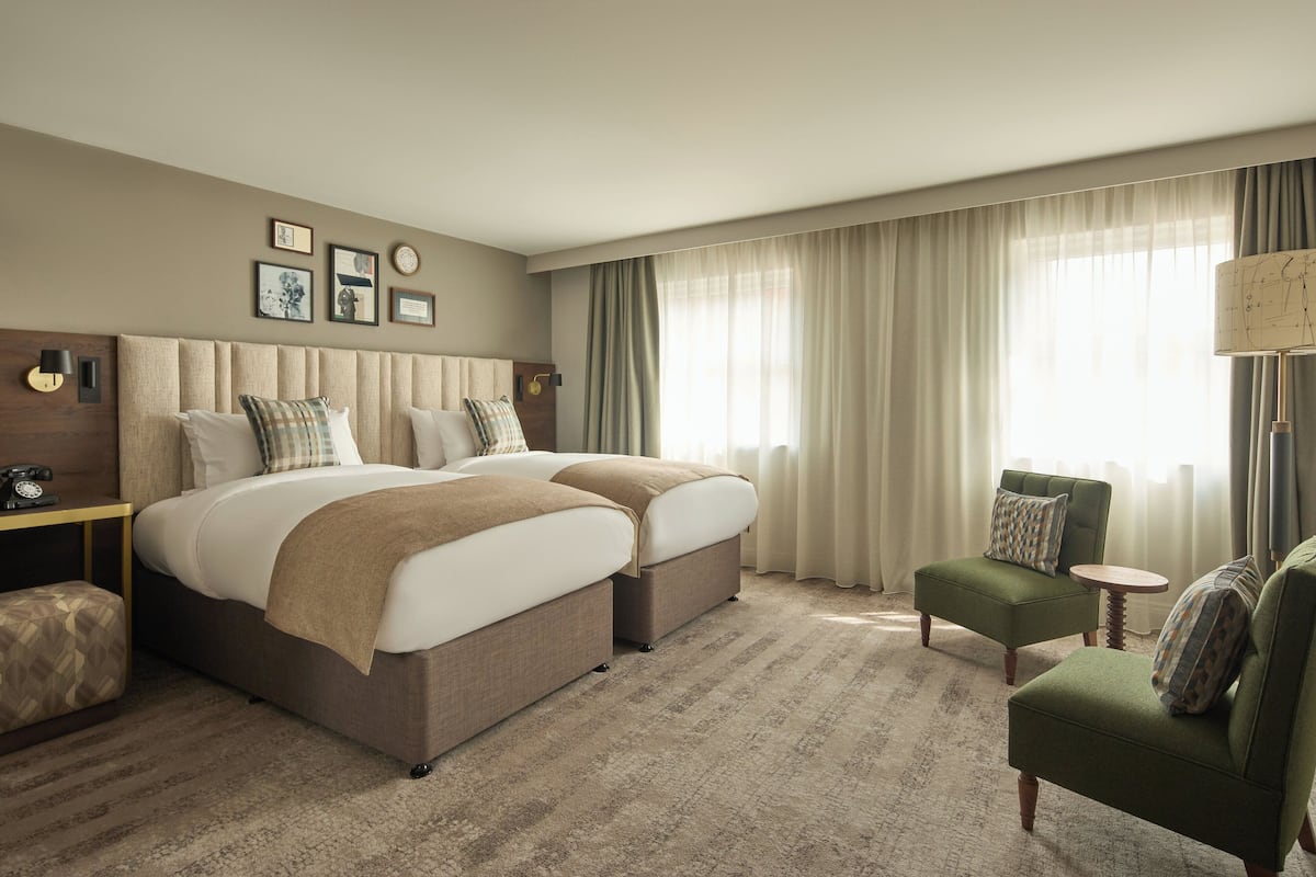 Habitación de hotel con dos camas, un sillón verde, una mesita redonda y una lámpara.