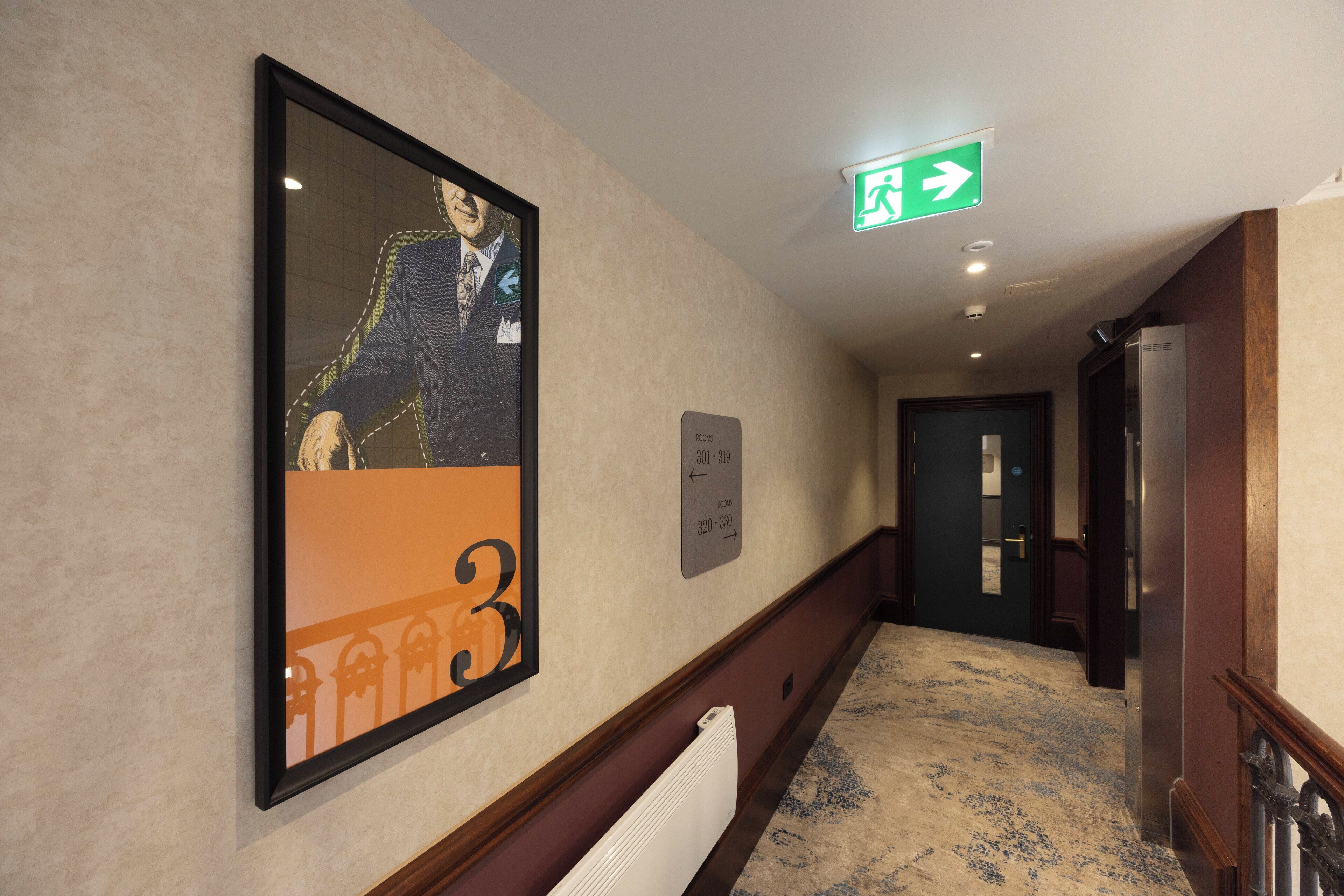 A hotel hallway with a framed portrait, a directional sign, and a door.