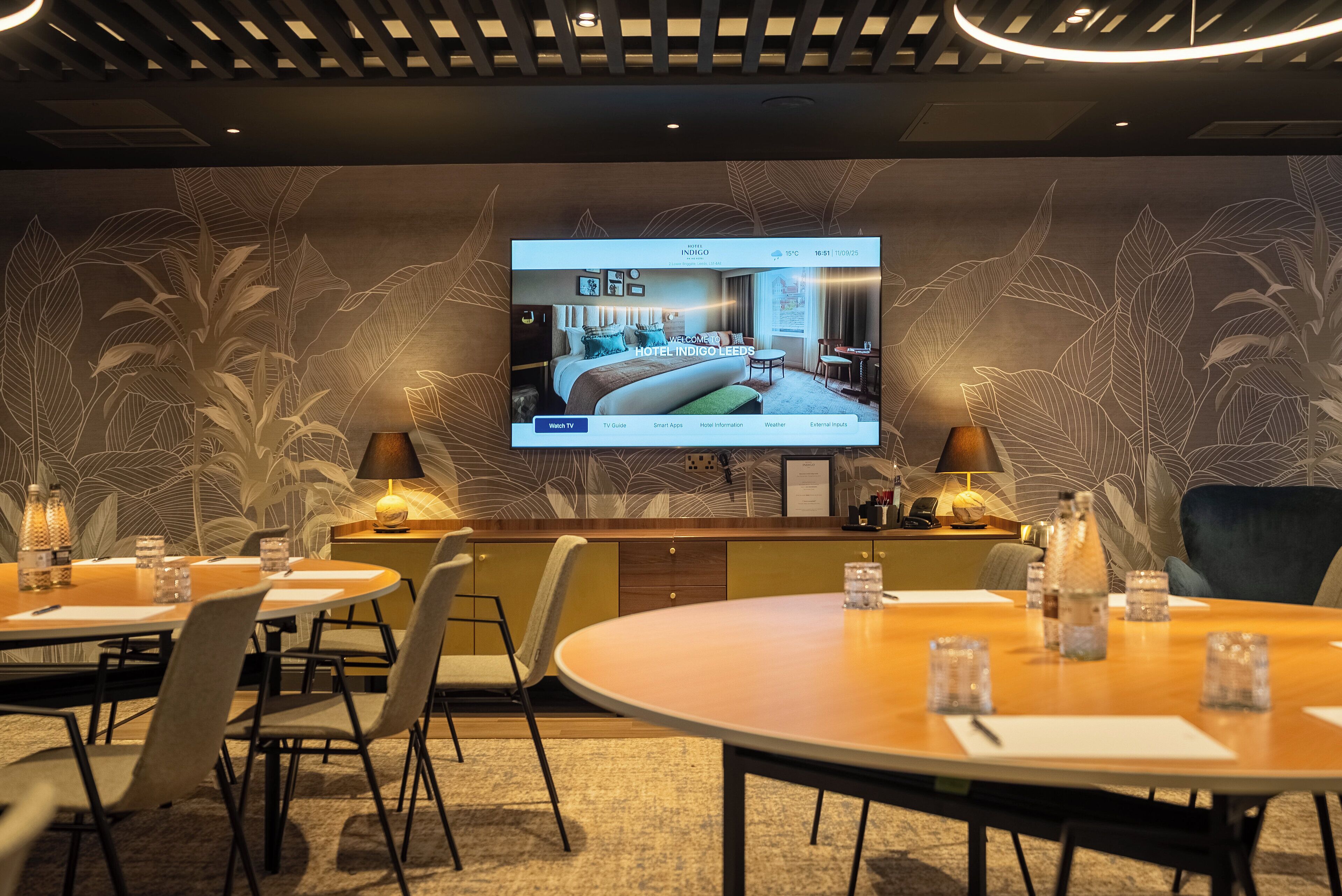 Ett hotellrum med en tv-skärm som visar en planritning över rummet, en bardisk och en matplats med stolar och bord.