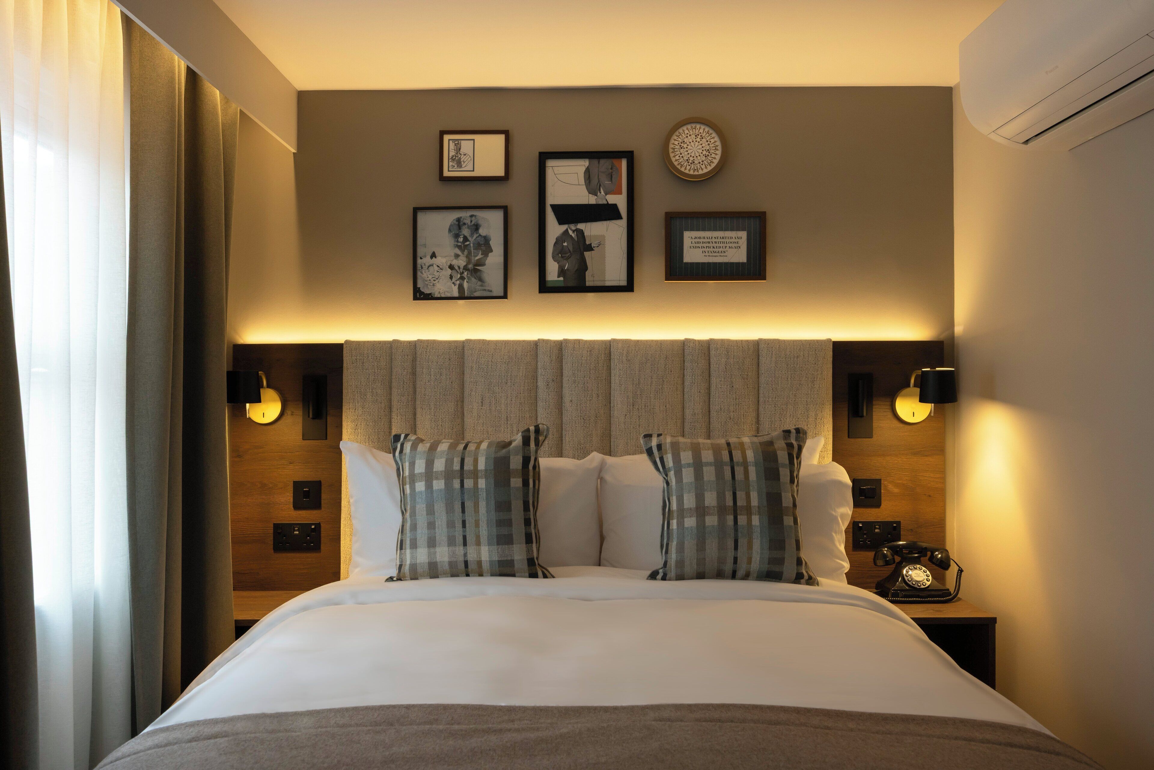 Una cama bien hecha con almohadillas escocesas, un cabecero con luces fijadas a la pared y cuadros en la pared.