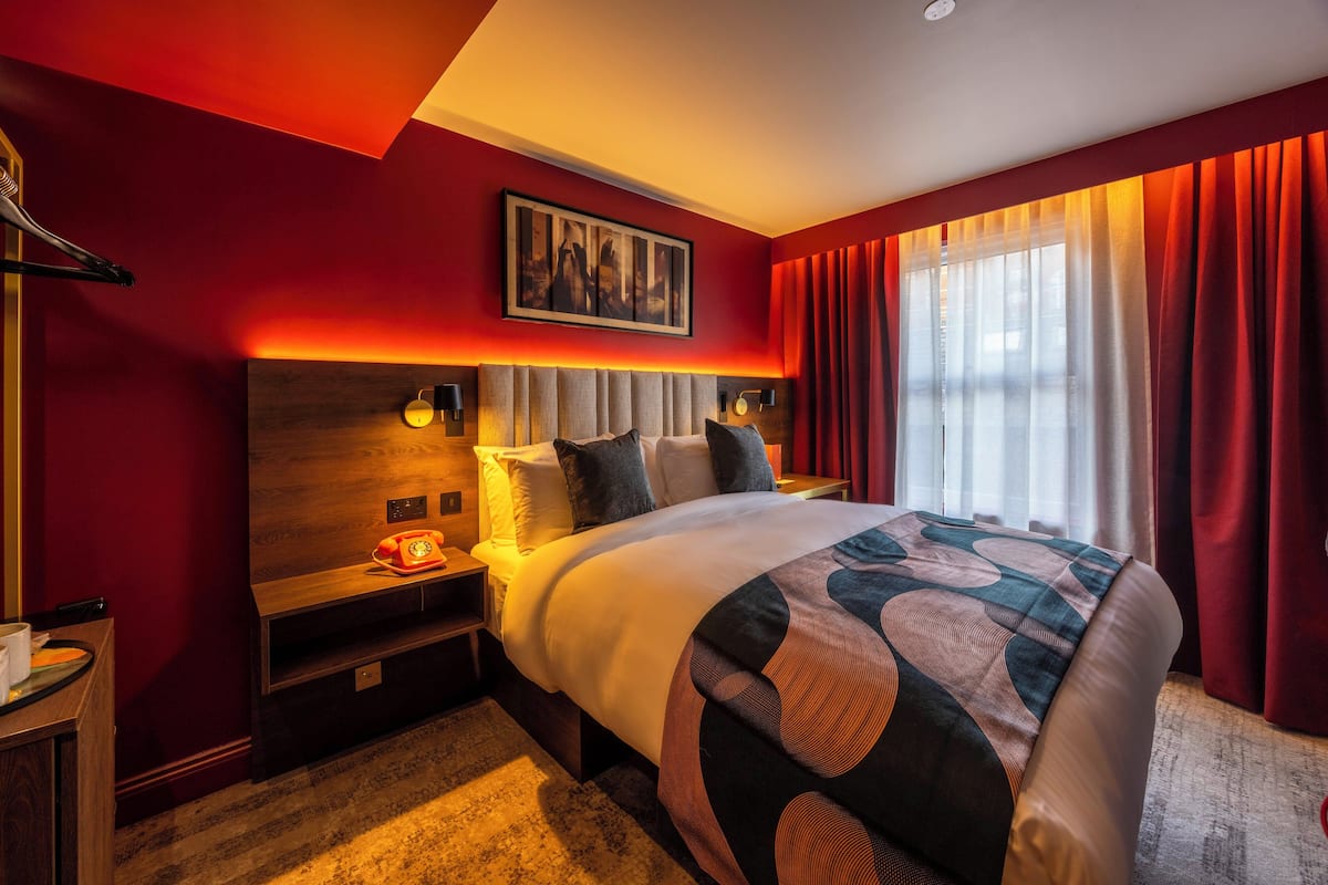 Una habitación de hotel moderna con una cama grande, una mesita de noche con teléfono, una obra de arte en la pared y una ventana con cortinas rojas.